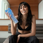 A woman with long dark hair, wearing a black bra and dark pants, is sitting on a sofa and smiling at the camera. She is holding a blue cylindrical Fleshlight Turbo Core Masturbator - Blue Ice in her right hand. The background shows a modern interior with wooden elements.